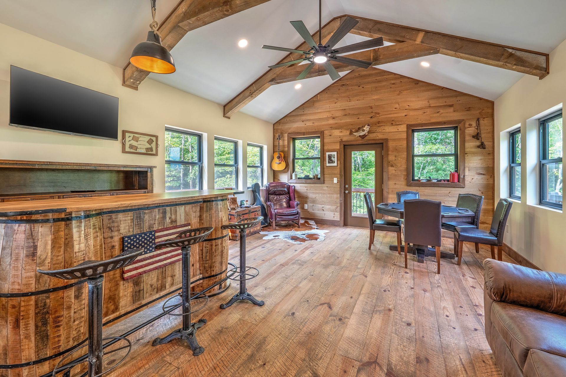 Rustic cabin living room with wood beams, fireplace, windows, dining table, and leather chair