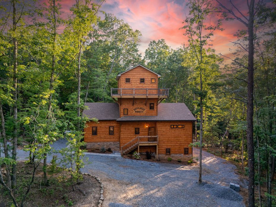 Wooden cabin in a forest clearing at sunset, framed by tall trees and a pink-orange sky.