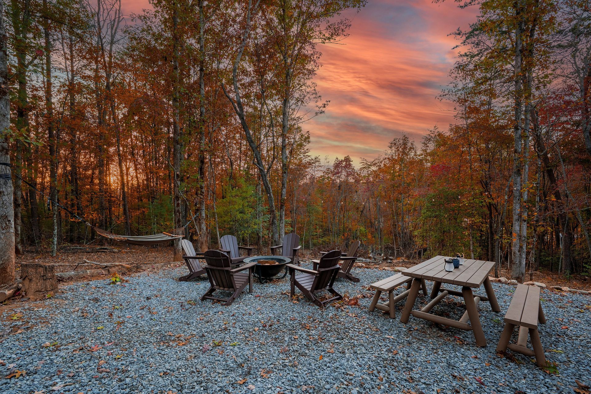 Outdoor fire pit with Adirondack chairs and picnic table at sunset in a wooded clearing