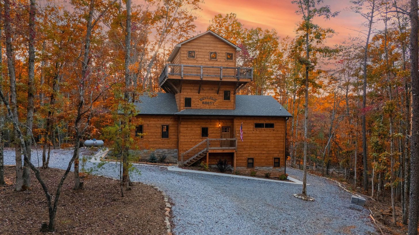 Rustic wooden cabin in autumn woods at sunset, with a gravel path and orange sky.