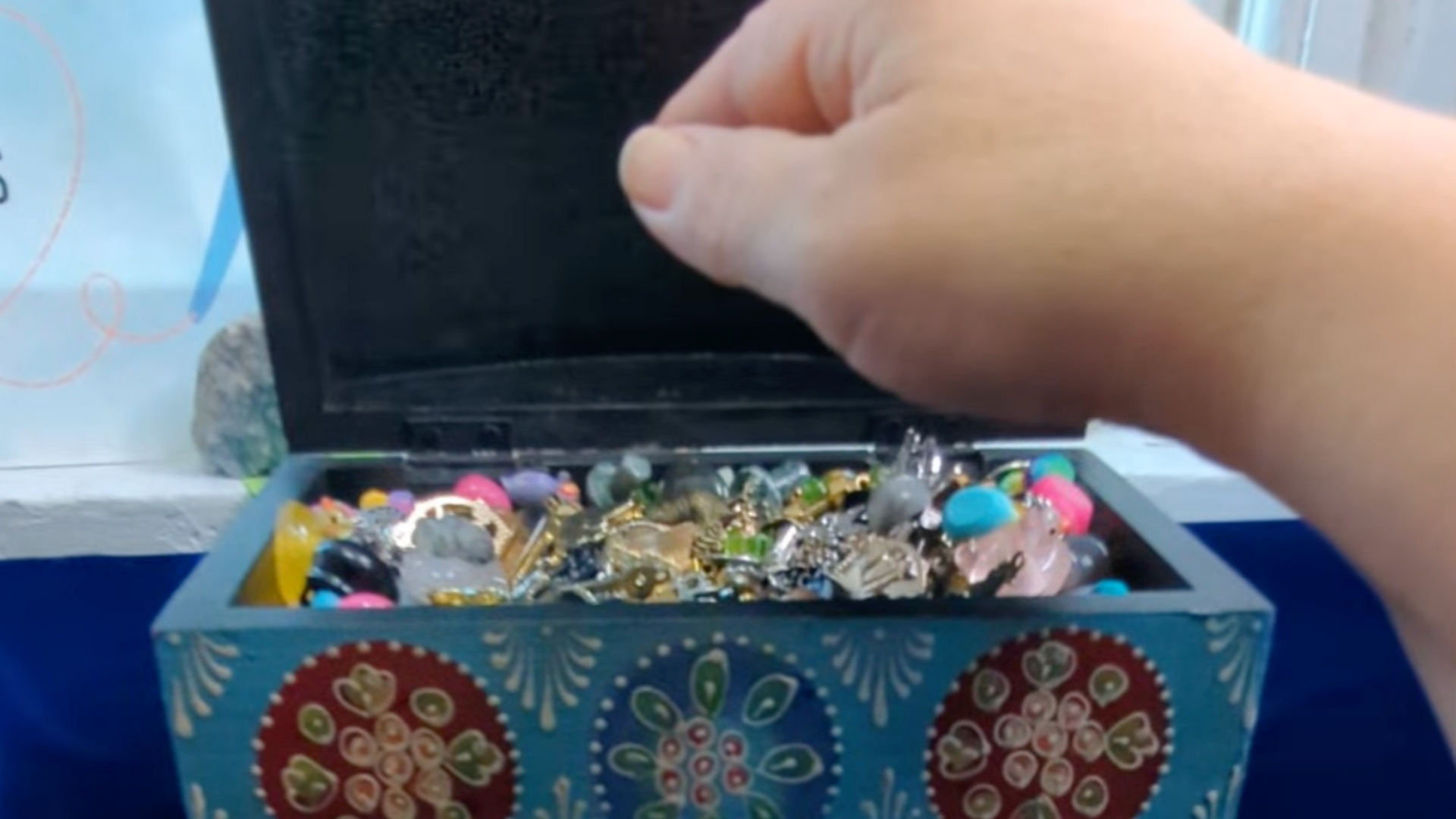 Hand opening a blue decorated chest filled with tiny charms of many colors, sizes, and shapes.