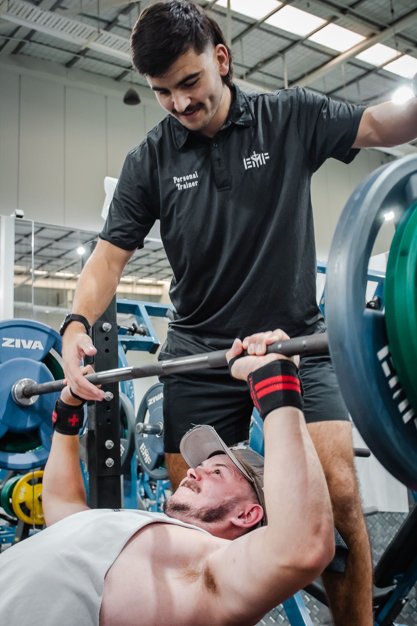 A gym trainer spots a person performing a bench press with heavy weights in a fitness facility.