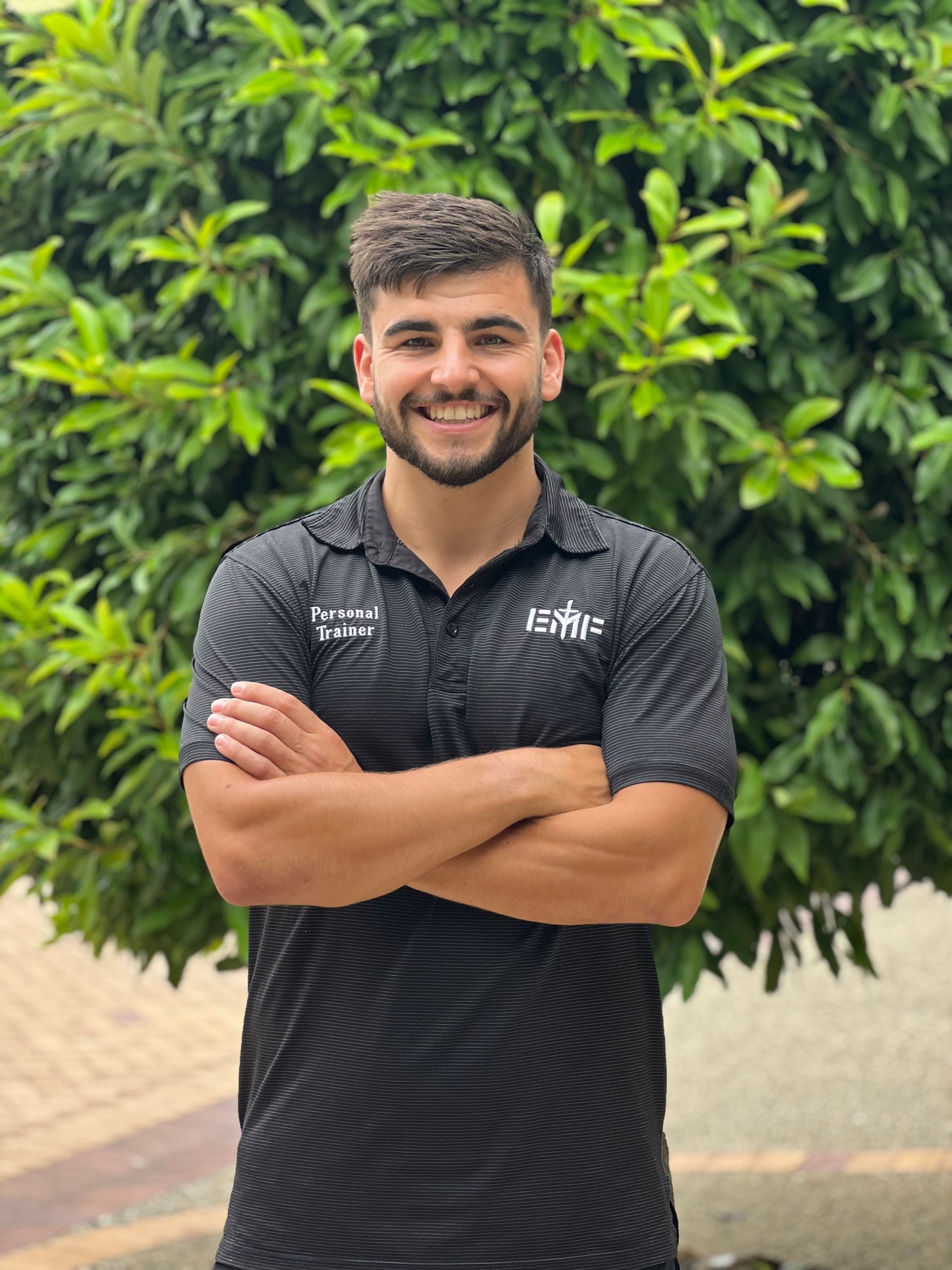A person with a beard, wearing a black patterned polo shirt, smiling with arms crossed in front of a green leafy tree.