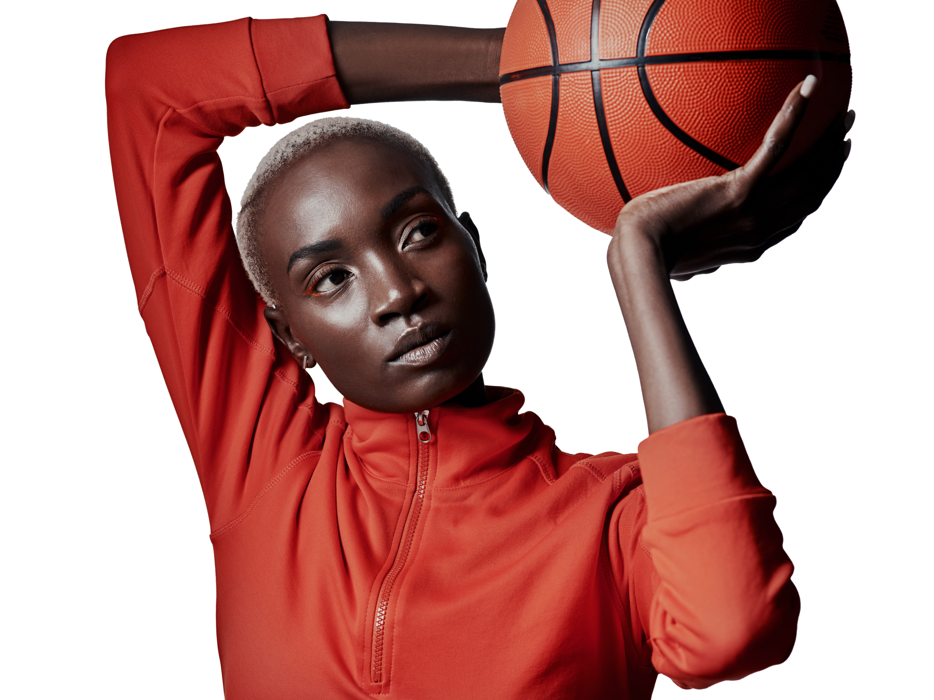 A person with short hair, wearing a bright orange top and black shorts, holds a basketball near their face against a beige.