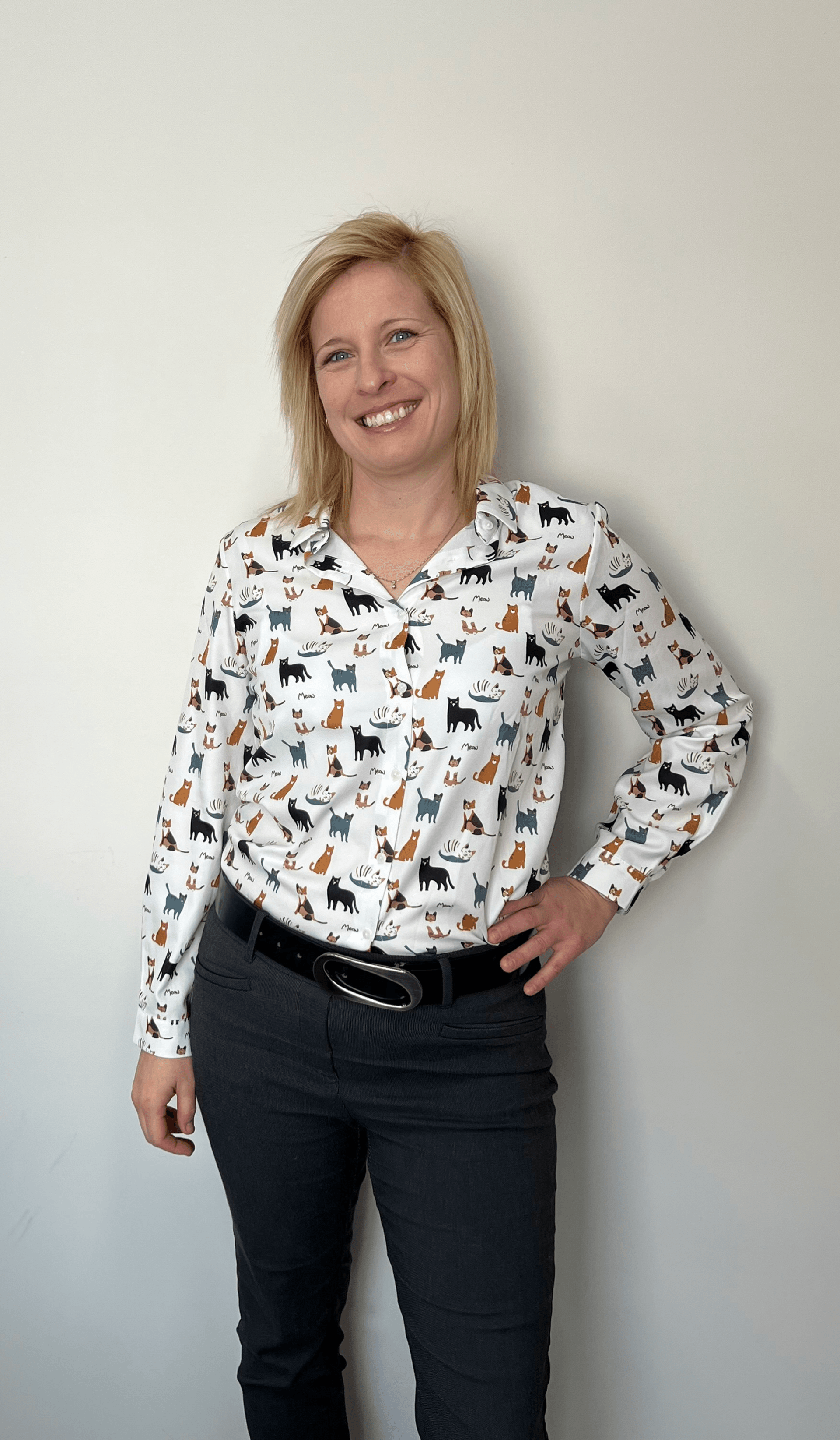 Une femme portant une chemise blanche avec des chats dessus se tient devant un mur blanc.