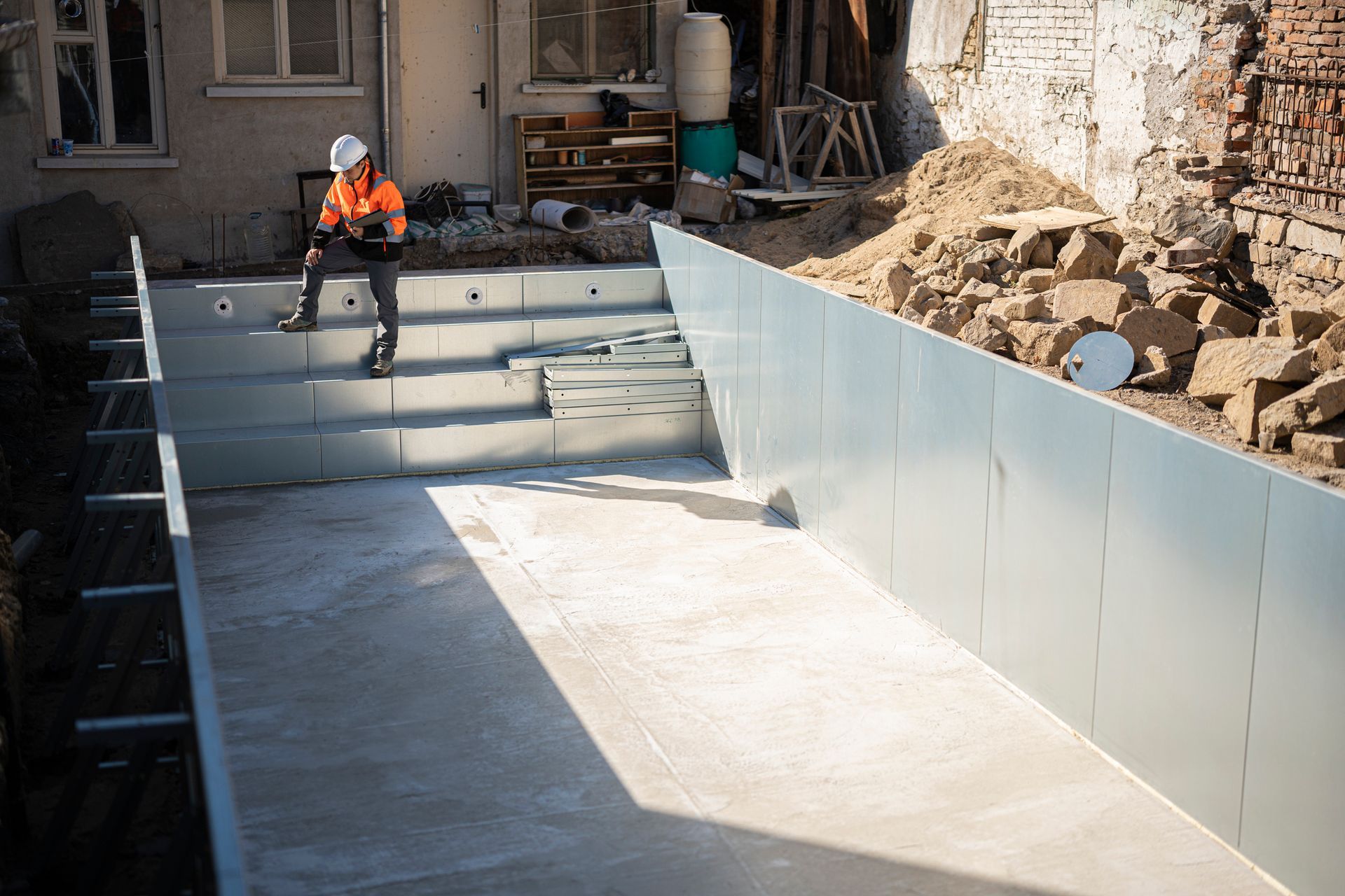 Construction worker in orange vest sits on edge of empty swimming pool.