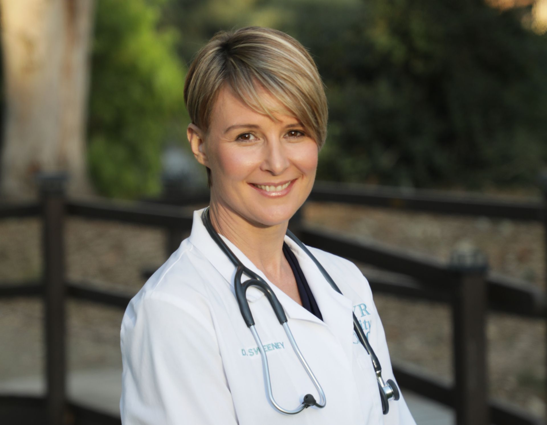 Woman wearing a white coat with stethoscope, smiling outdoors.