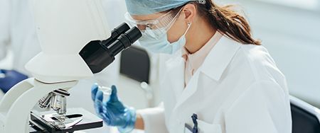 A person in a lab coat, goggles, and mask looks into a microscope while holding a small glass container.
