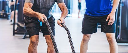 Two men in a gym exercising with battle ropes. One wears a gray shirt, the other a blue.