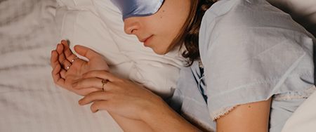 A person asleep in bed, wearing a sleep mask, hands folded, ring visible.