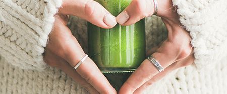 Hands forming a heart around a green smoothie in a glass. The person wears rings and a white sweater.