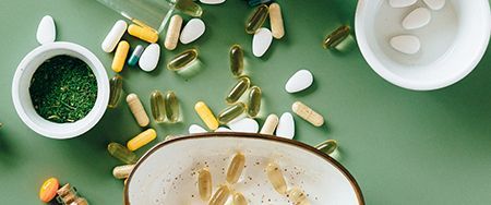 Overhead shot of various pills and supplements in small white bowls on a green surface.