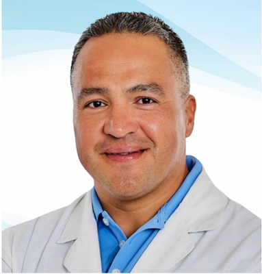 Man in white coat and blue shirt smiles at the camera against a light blue background.
