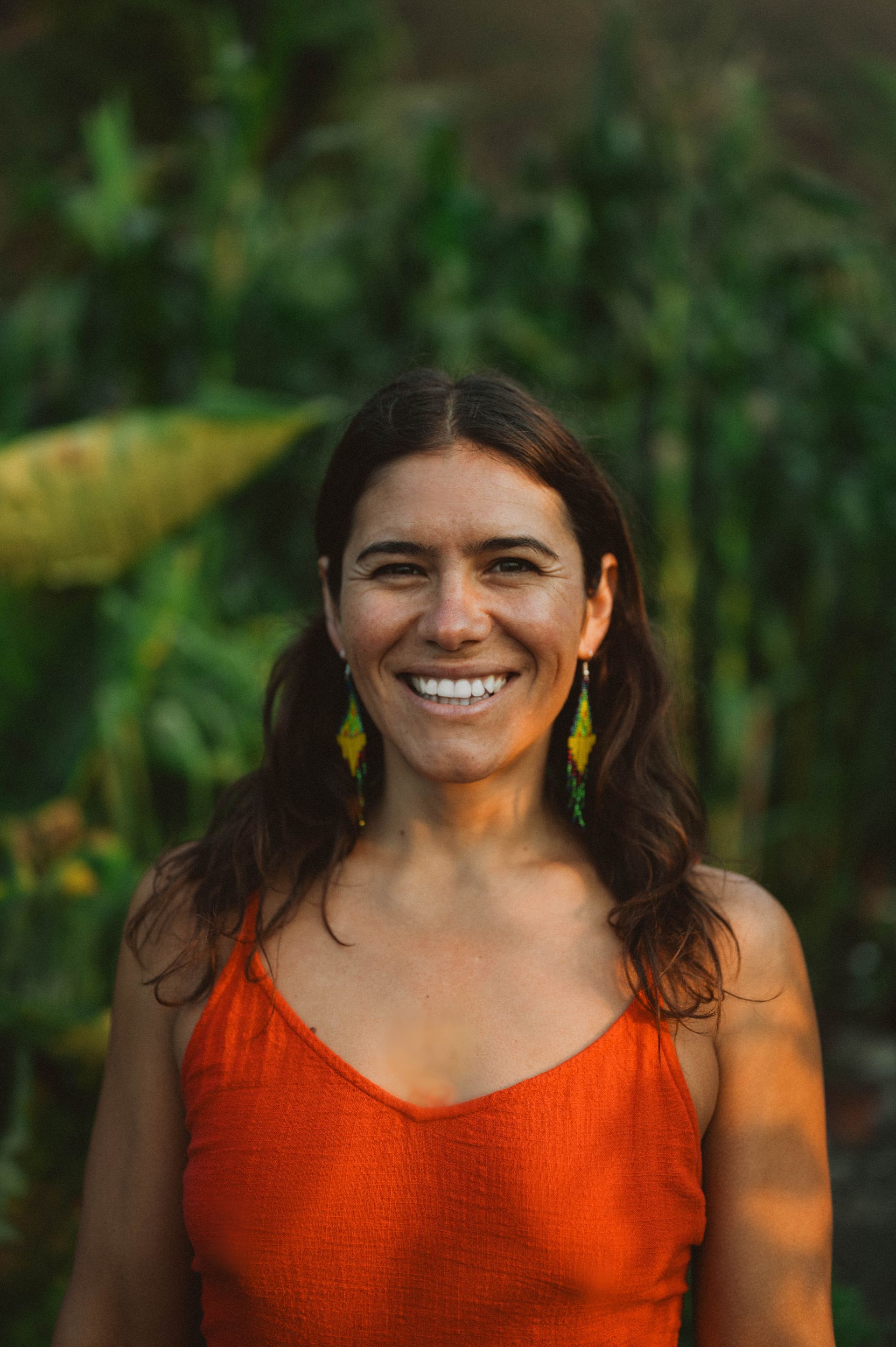 Woman smiling outdoors, wearing orange top, green leafy background, dangling earrings.