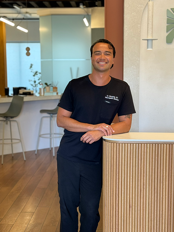 Man in black t-shirt and pants smiles, leaning on a counter in a modern office.