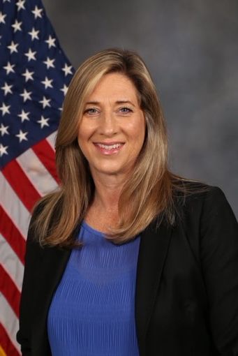 Woman smiling, in a blue shirt and black blazer, with the American flag in the background.