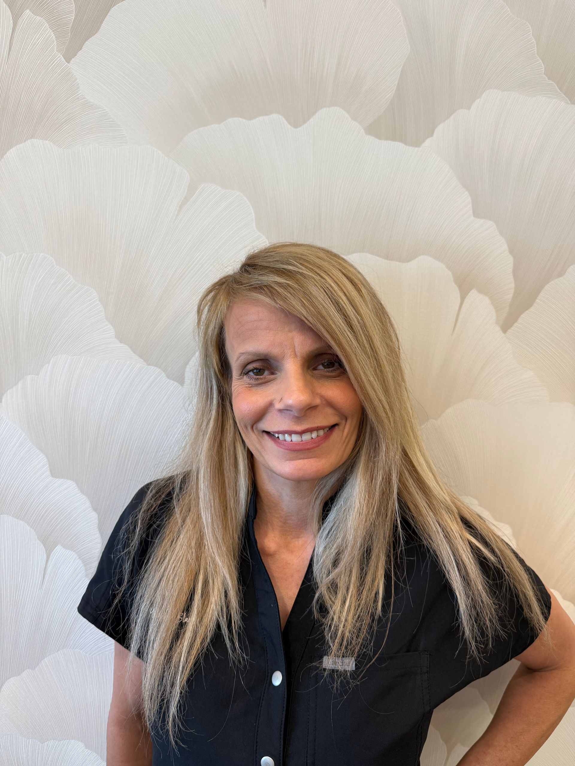 Woman smiling outdoors, wearing orange top, green leafy background, dangling earrings.