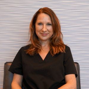 Woman smiling outdoors, wearing orange top, green leafy background, dangling earrings.