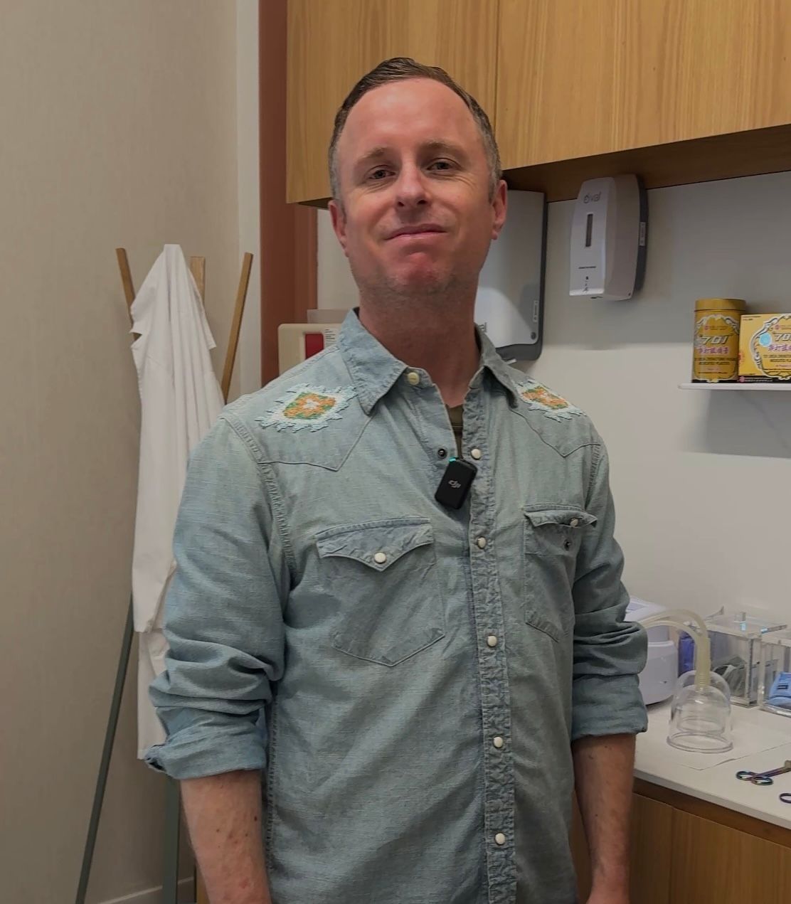 Man in denim shirt standing indoors, looking at the camera.