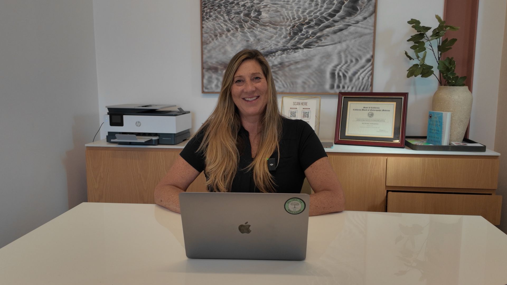 Woman smiling, in a blue shirt and black blazer, with the American flag in the background.