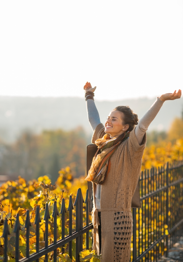 Woman with arms raised, smiling near a fence, overlooking a fall landscape with golden foliage.