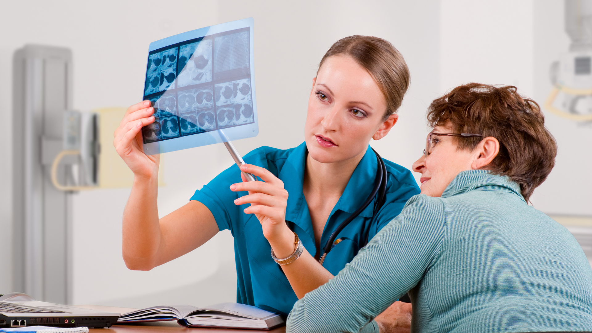 Doctor pointing at X-ray, explaining to a patient. Blue medical scrubs, examination room setting.