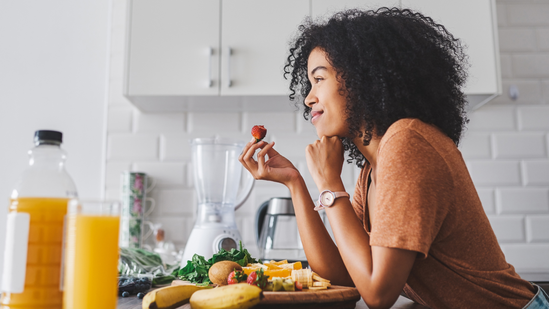 Woman in kitchen holding strawberry, smiling, with fruit and blender.