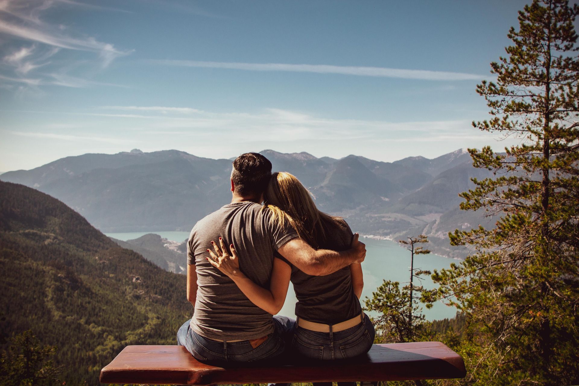 Couple with arms around each other, sitting on a bench overlooking mountains and a lake on a sunny day.