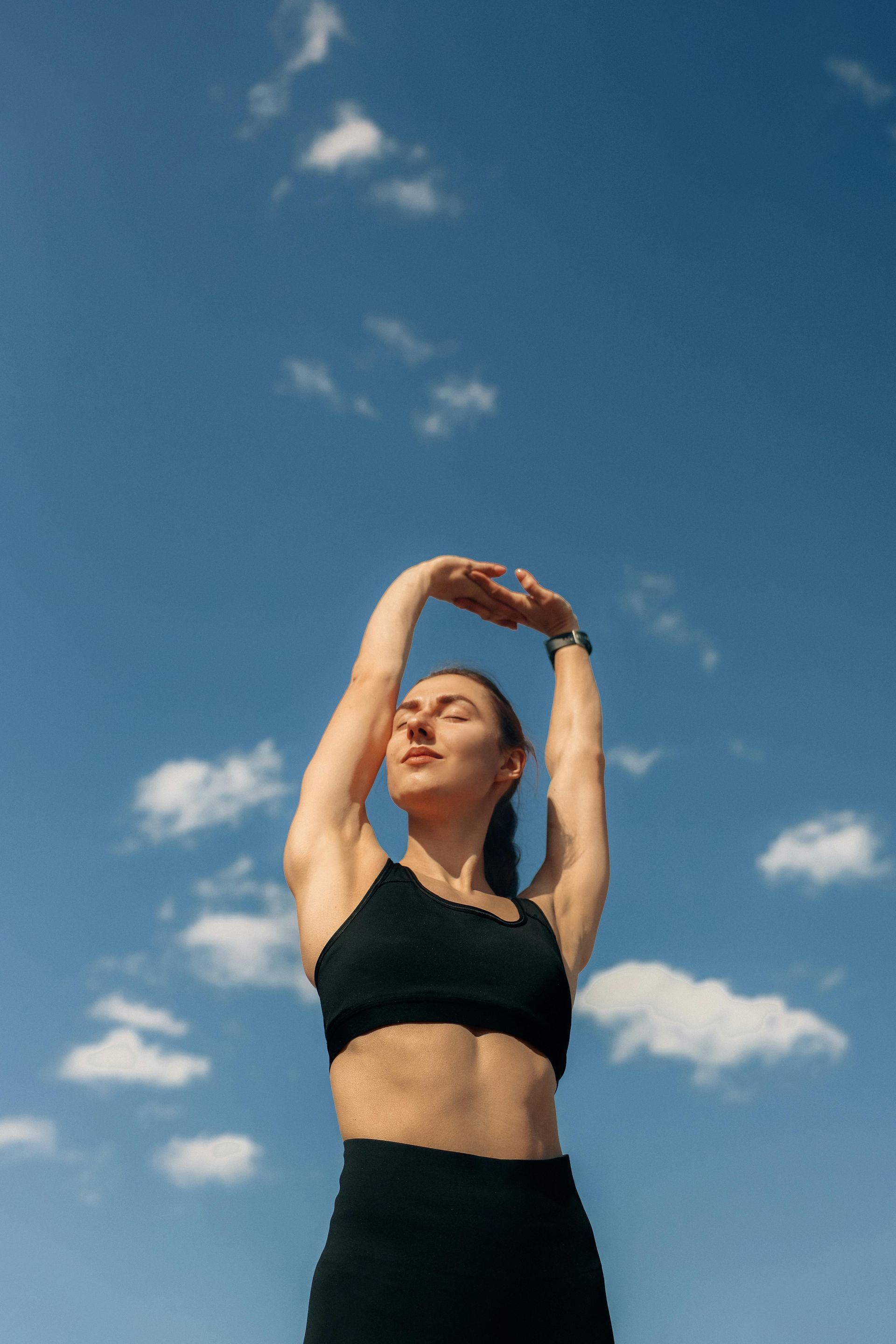 Woman in black sportswear stretching arms overhead against a blue sky with clouds.