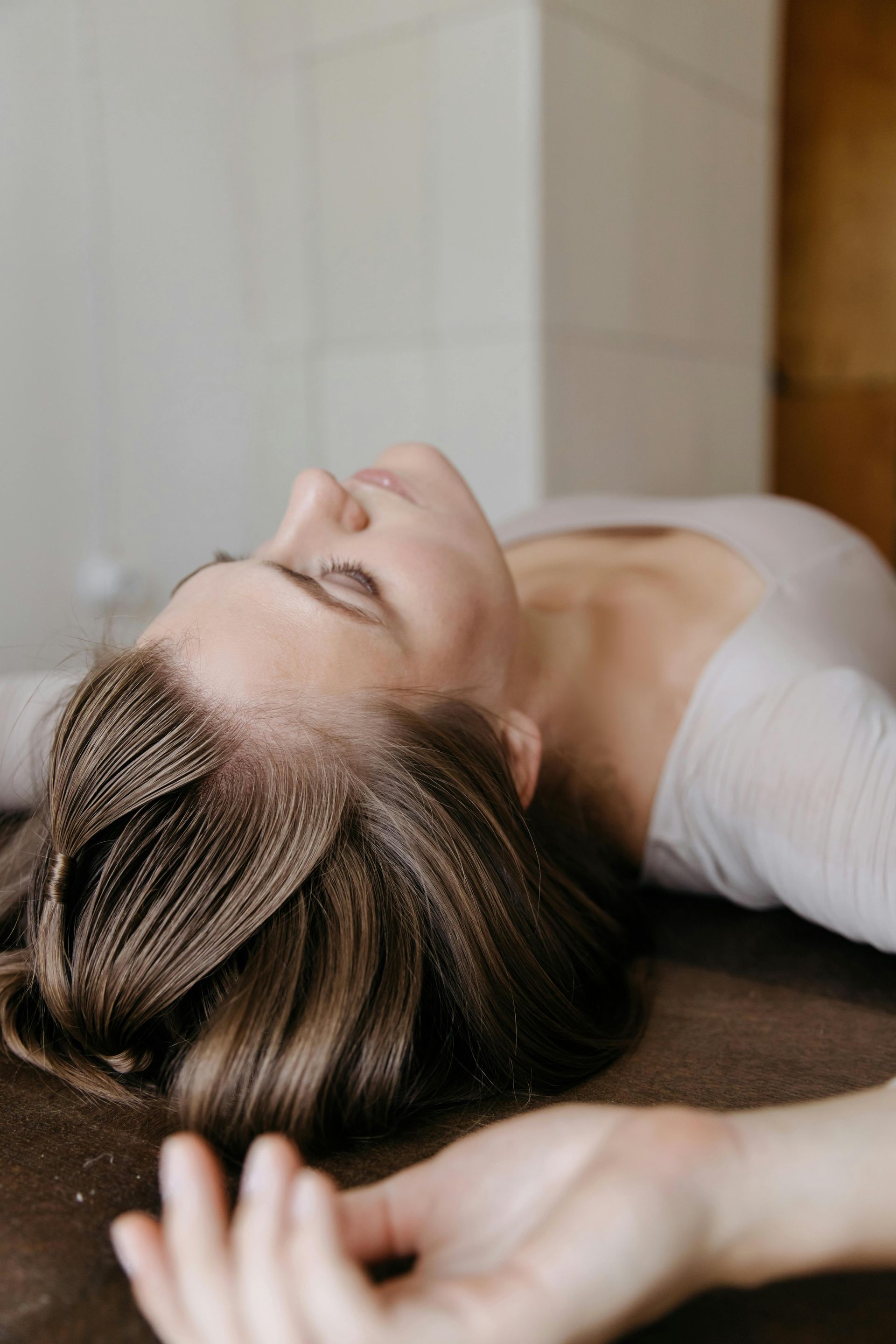Woman lying down, head tilted back, eyes closed, neutral expression. Arms outstretched, in a light-colored room.