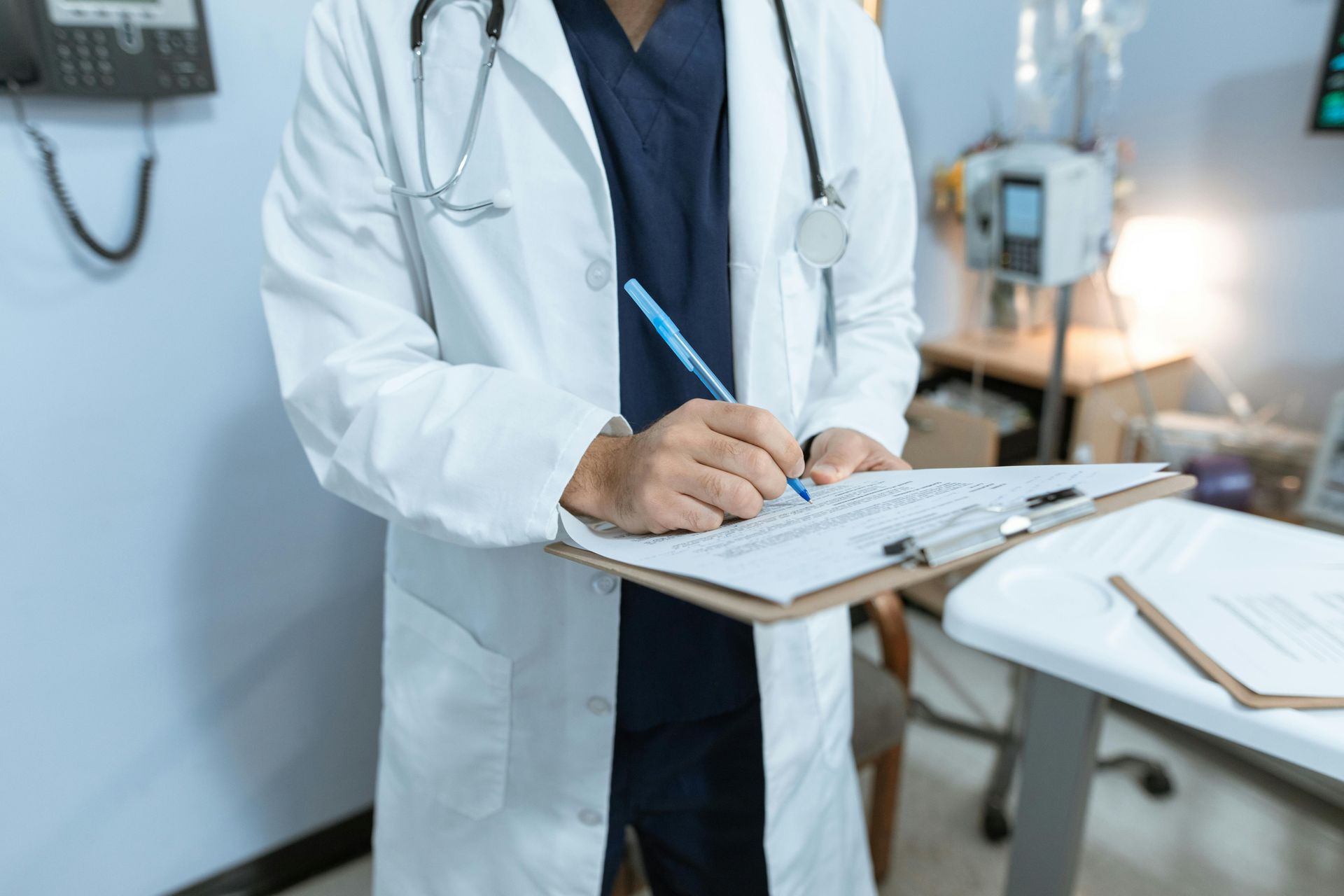 Doctor in white coat writing on a clipboard in a medical setting, stethoscope around their neck.