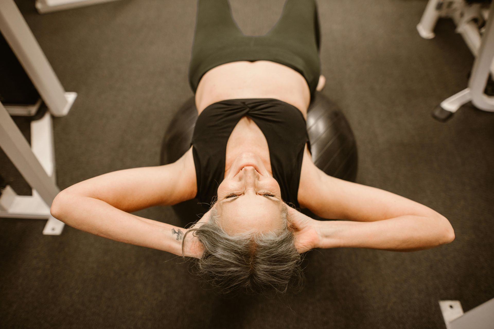 Woman doing crunches on a fitness ball in a gym. She is wearing black activewear, and smiling with hands behind her head.