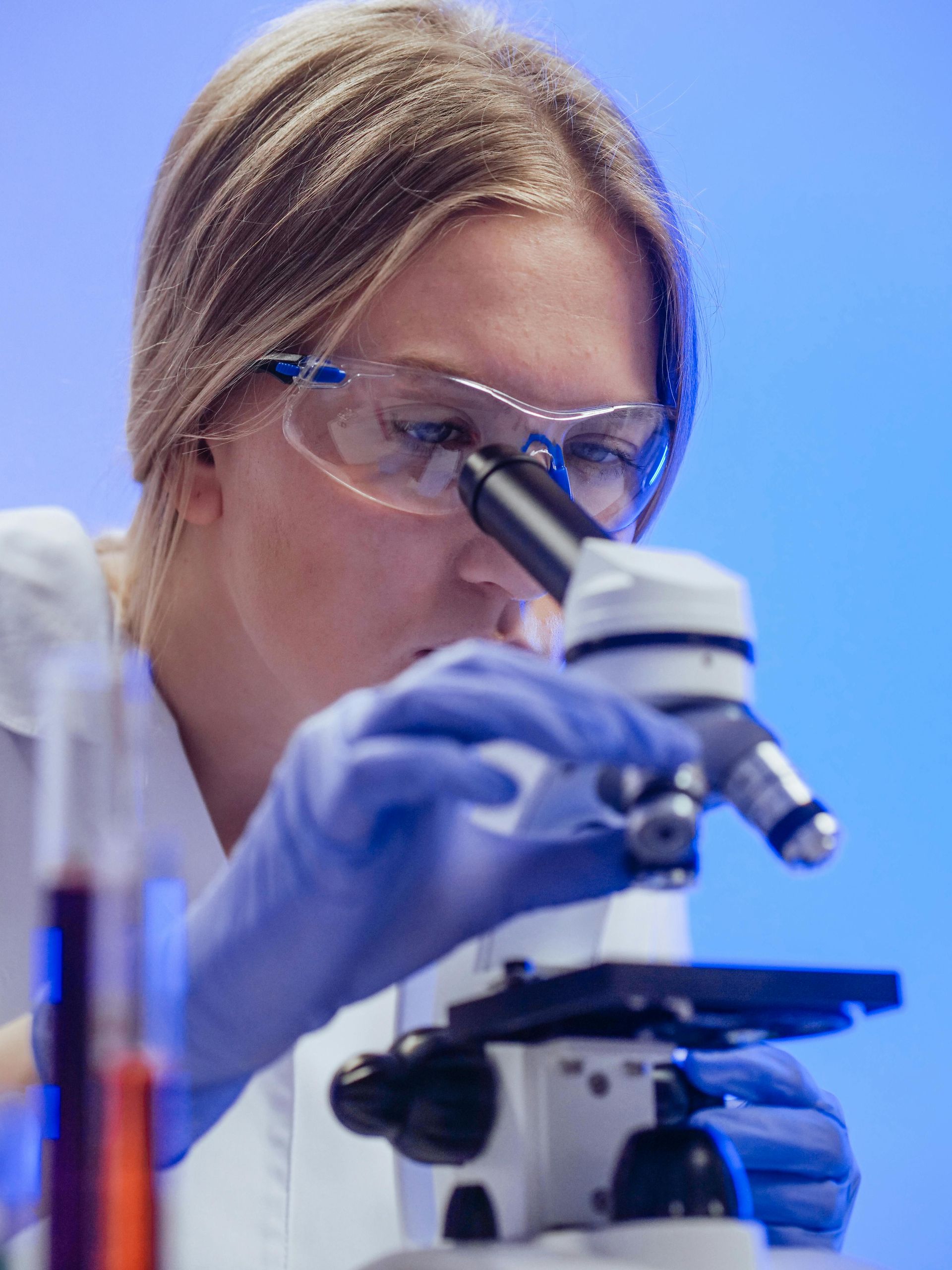 Scientist looking through a microscope, wearing safety glasses and gloves, with test tubes in the background.