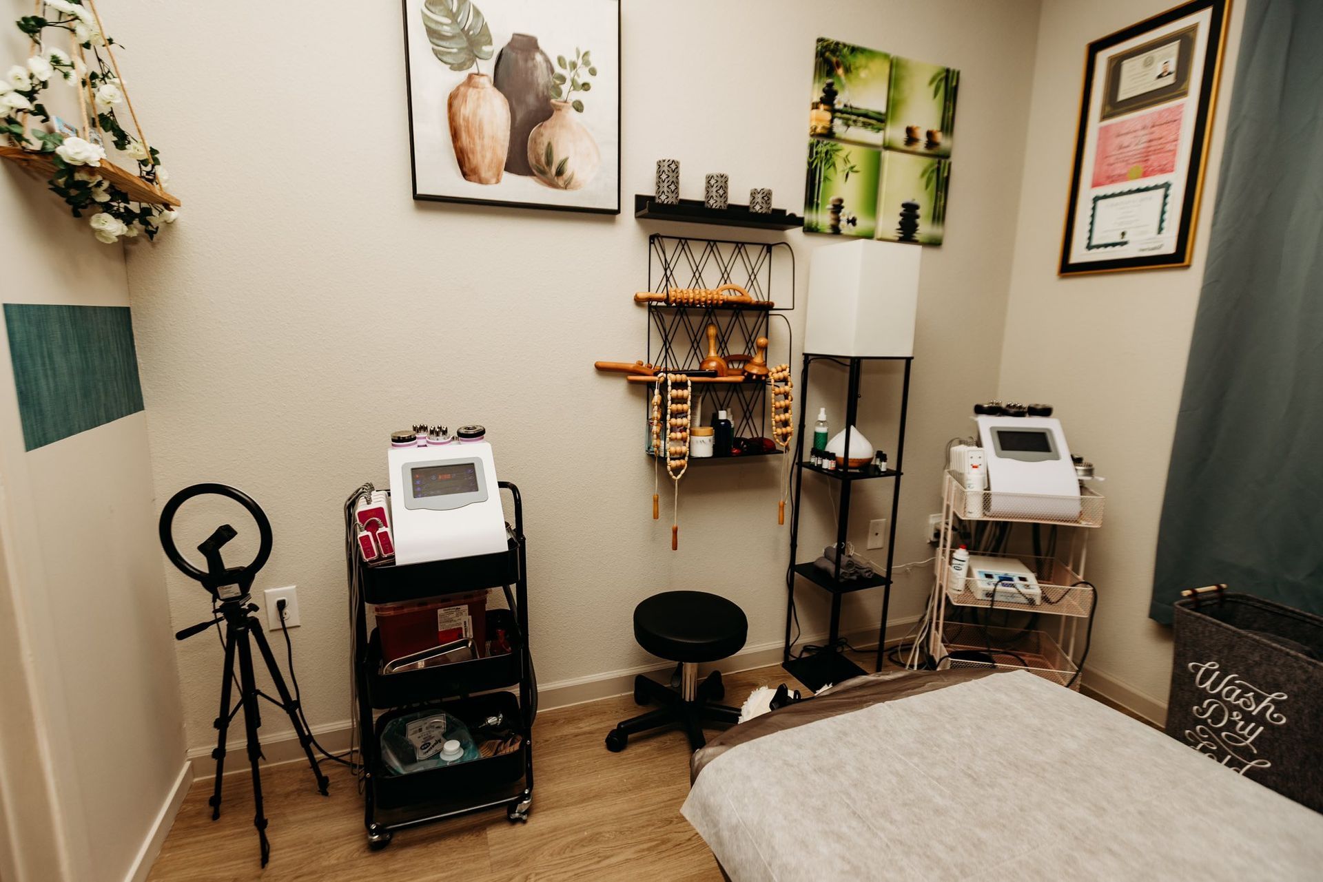 A treatment room with medical equipment, shelves, and a massage table. Light-colored walls and wood flooring.