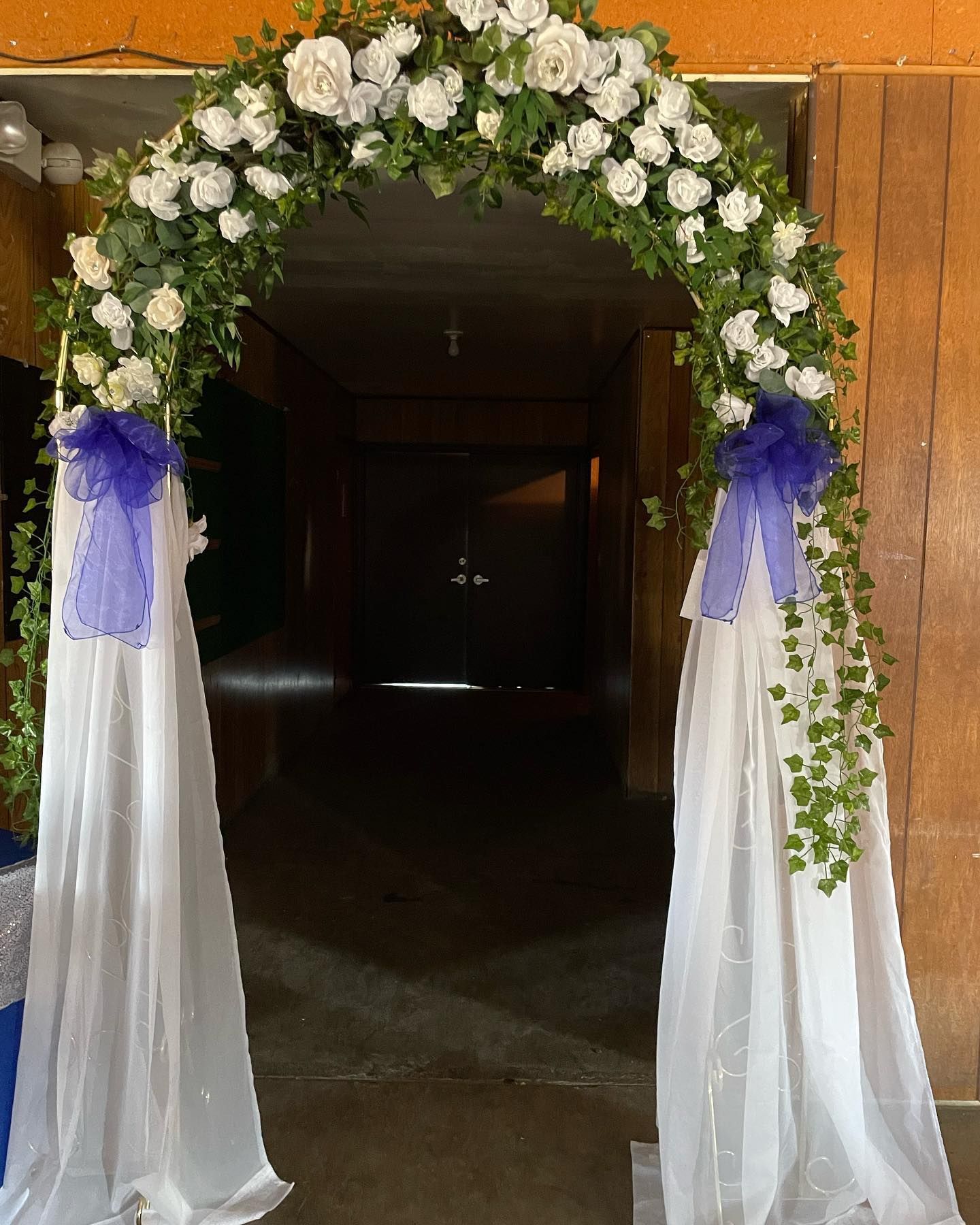 Floral archway with white roses, greenery, blue accents, and white fabric curtains.
