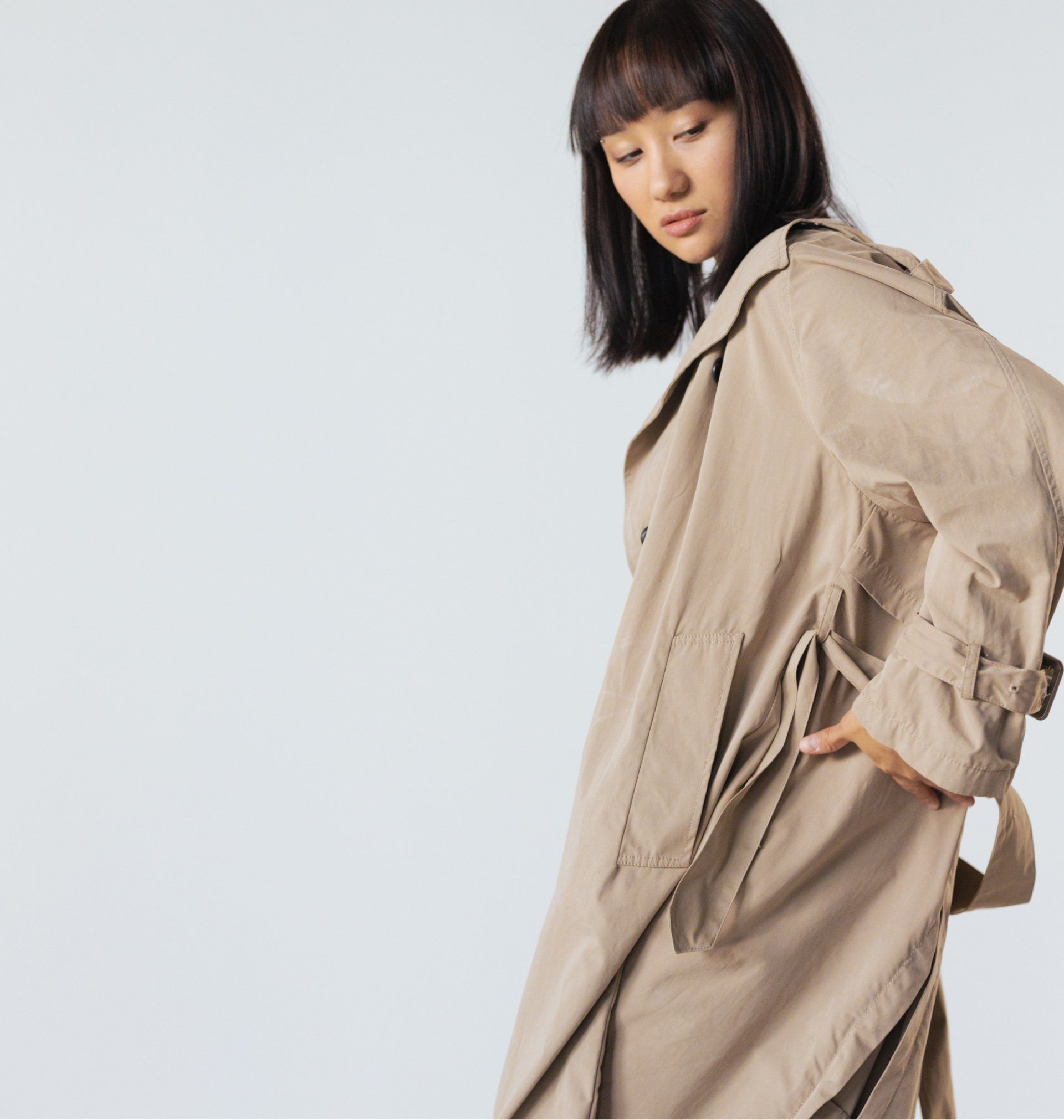 Woman in tan trench coat, looking down, hand in pocket, against a light background.