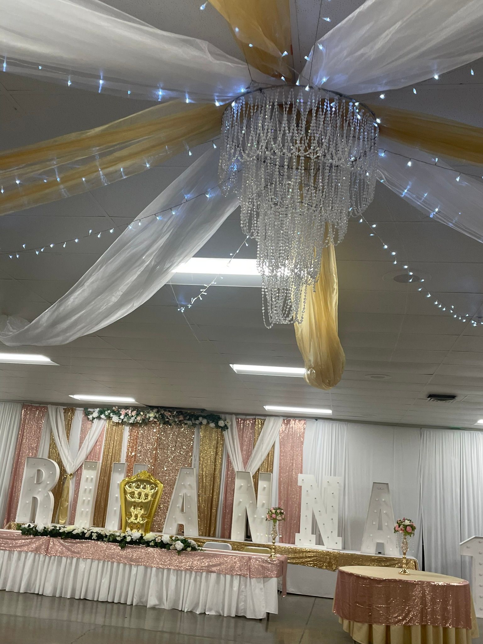 Event hall decorated with gold and white draping, chandelier, and a table with the name 