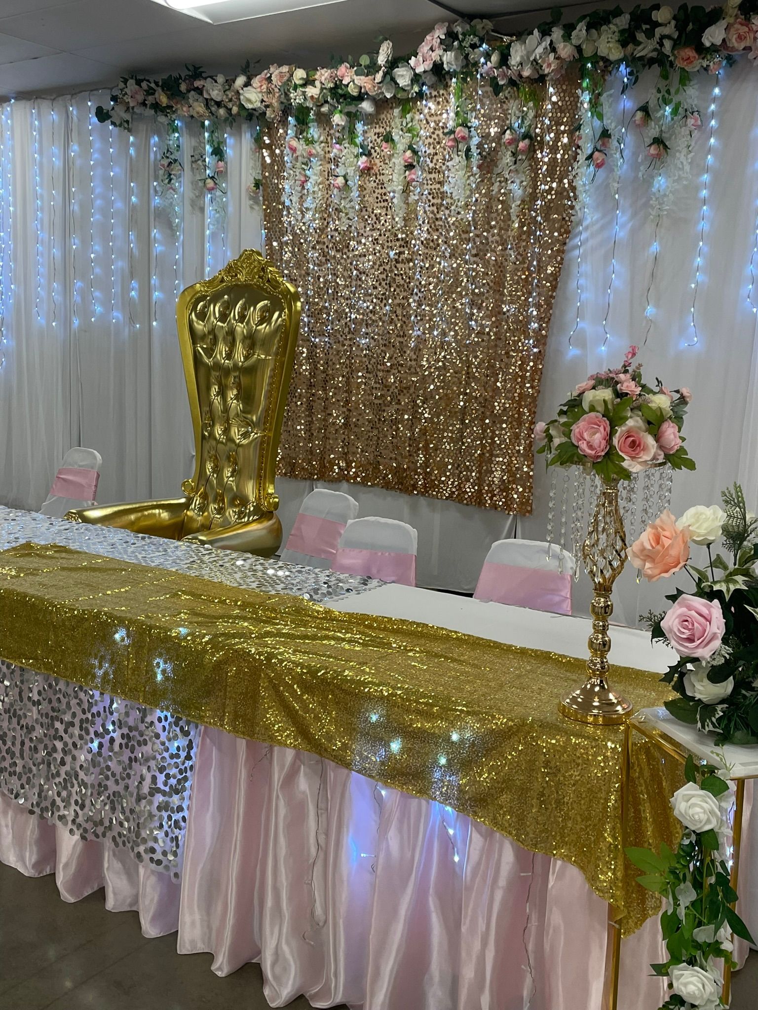 A decorated table with gold sequins and pink accents, backdrop of hanging lights and flowers.