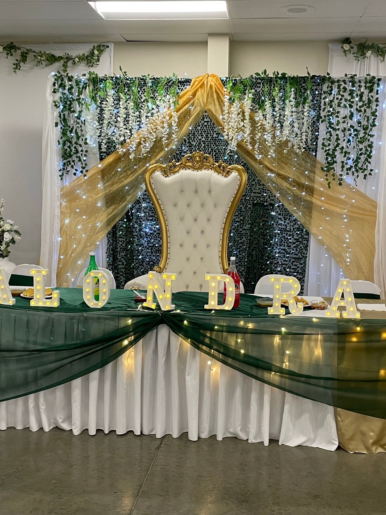 Elegant white and gold decorated party table with large throne chair, draping, and lights. 