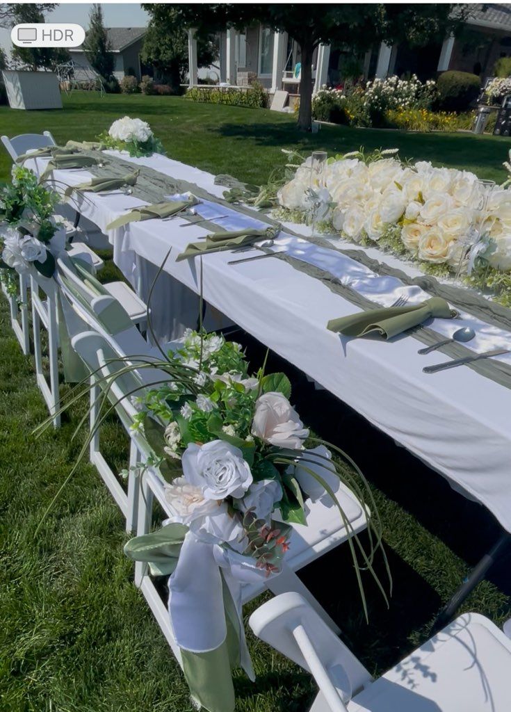 Long white table set up outdoors, decorated with white flowers and greenery, white chairs arranged along the sides.