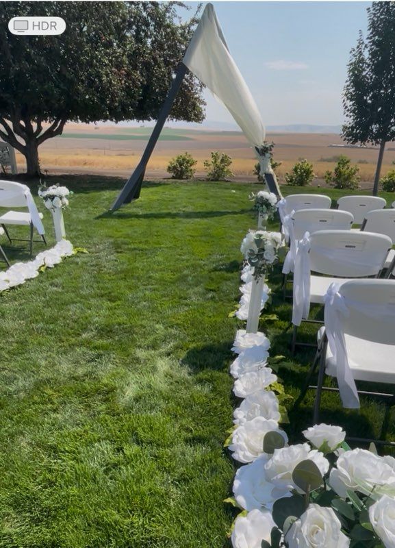 Wedding ceremony setup: white chairs, aisle with floral decorations, draped canopy, green grass, and scenic background.