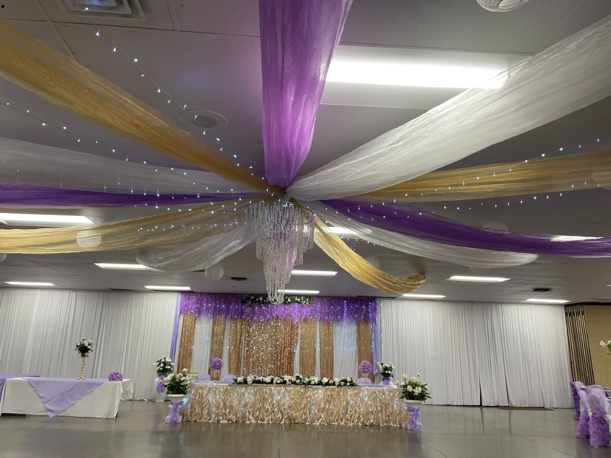Wedding reception hall decorated with purple, gold, and white fabric draping and lights.