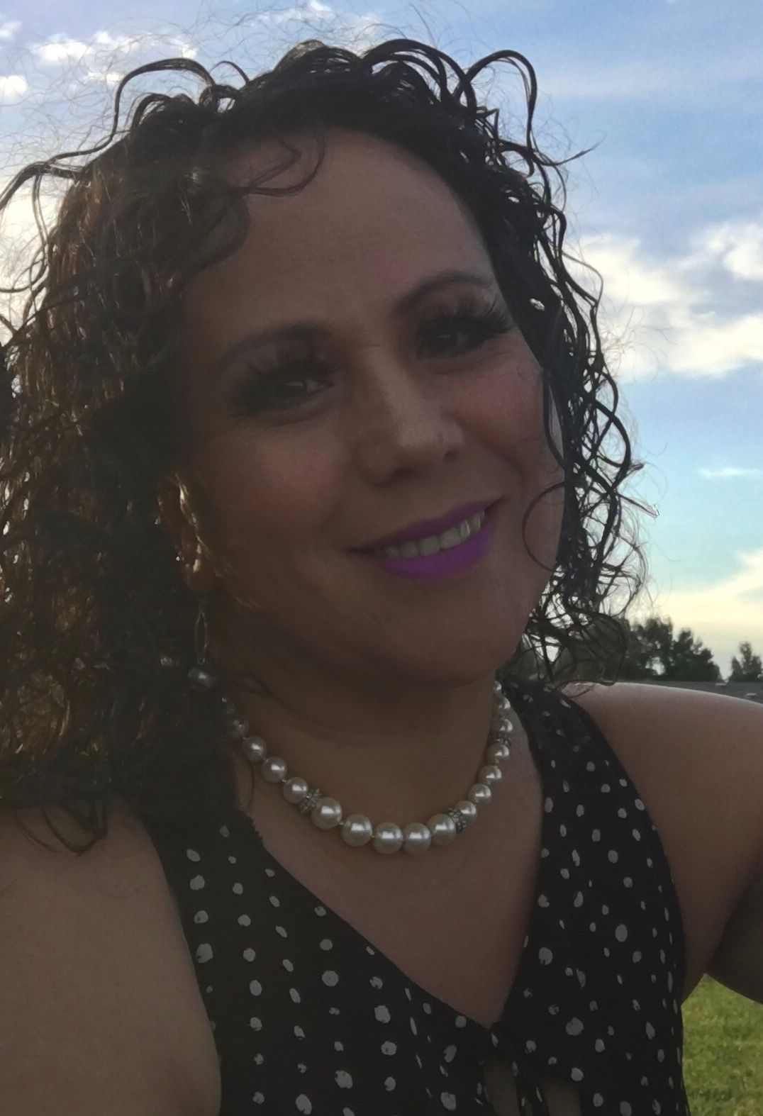 Woman with curly hair and a pearl necklace smiles, posing outdoors.