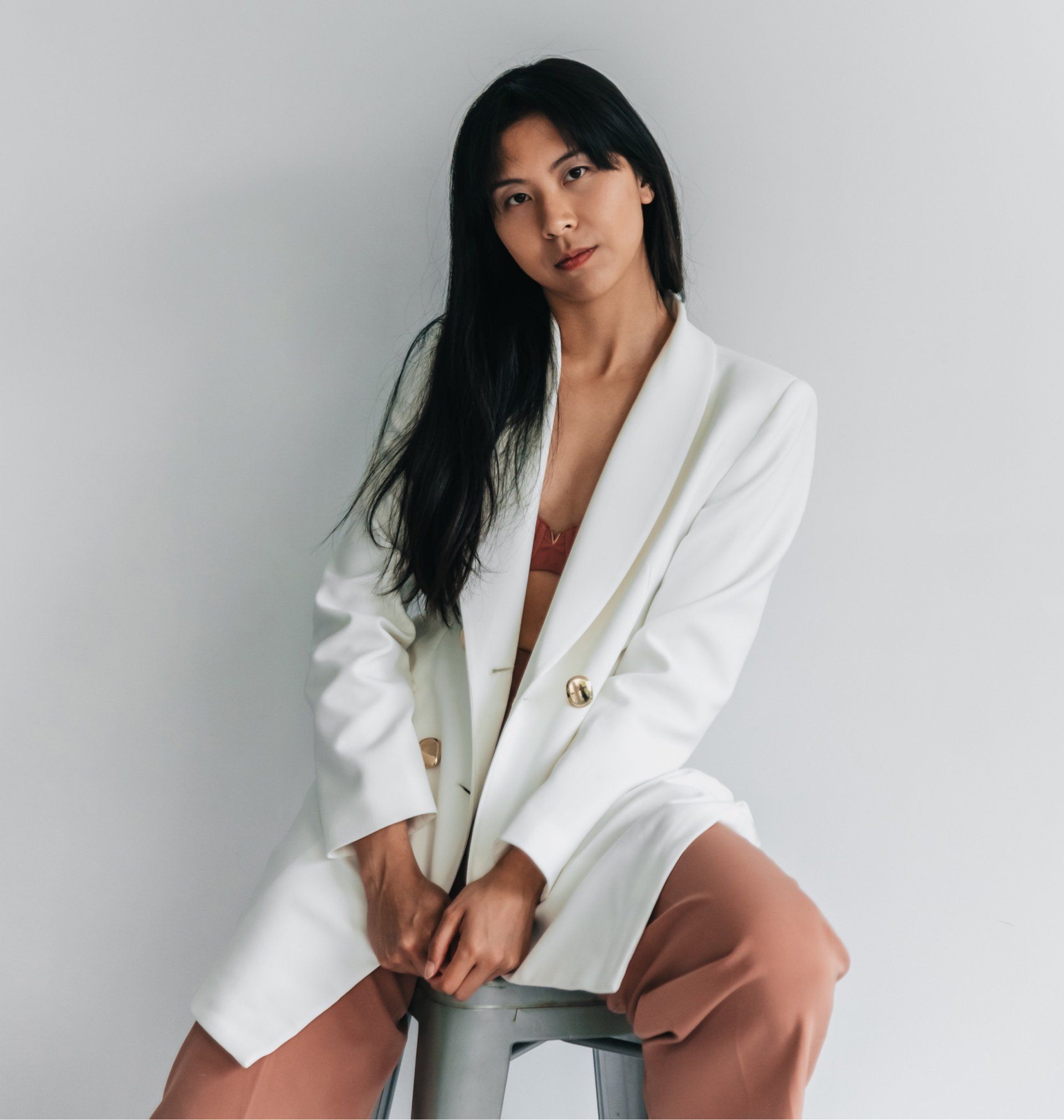 Woman in a white blazer and brown pants, leaning back with eyes closed against a white wall.