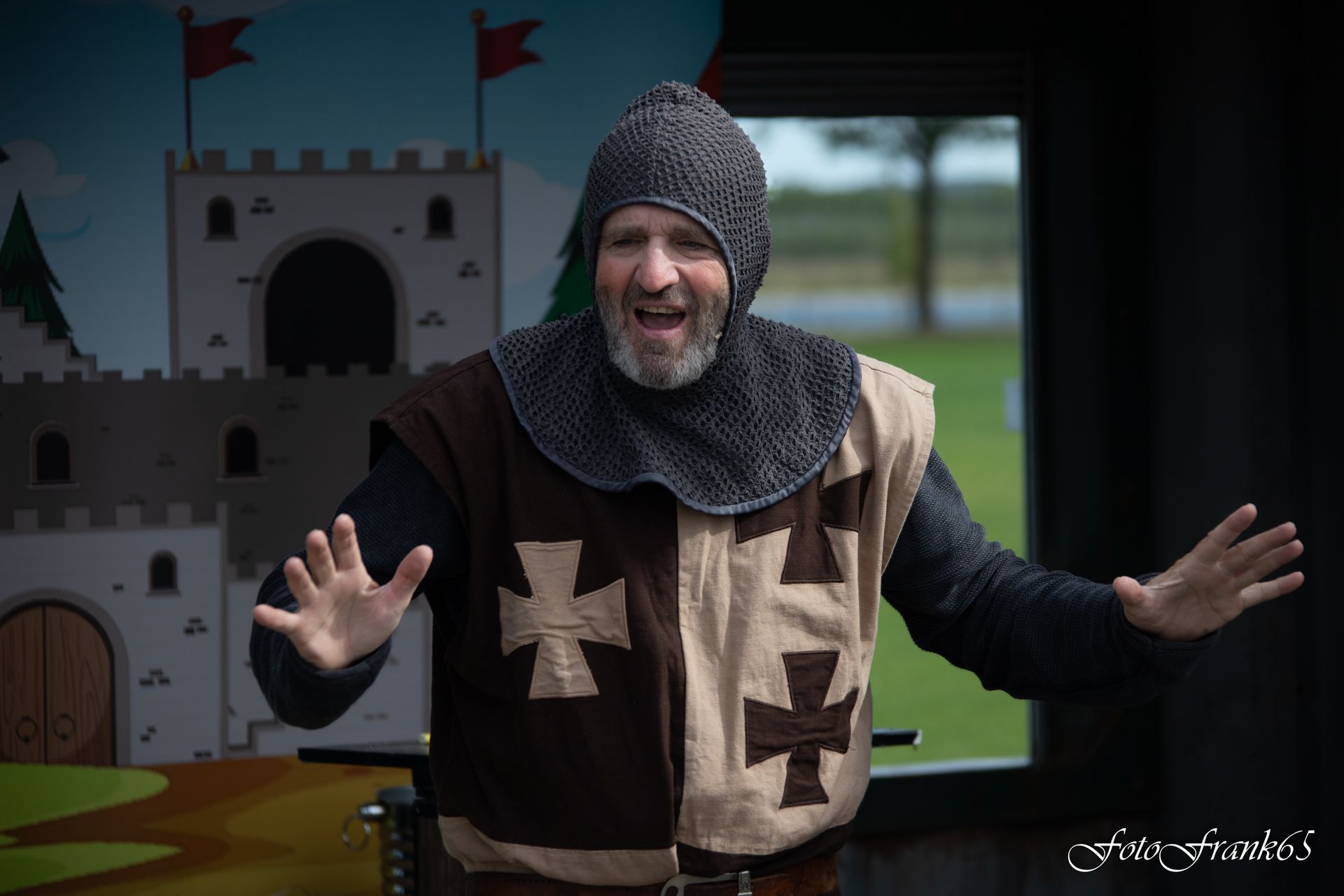 Een man in een ridderkostuum treedt op een podium op met een kasteel en bomen als achtergrond, tegenover een publiek.