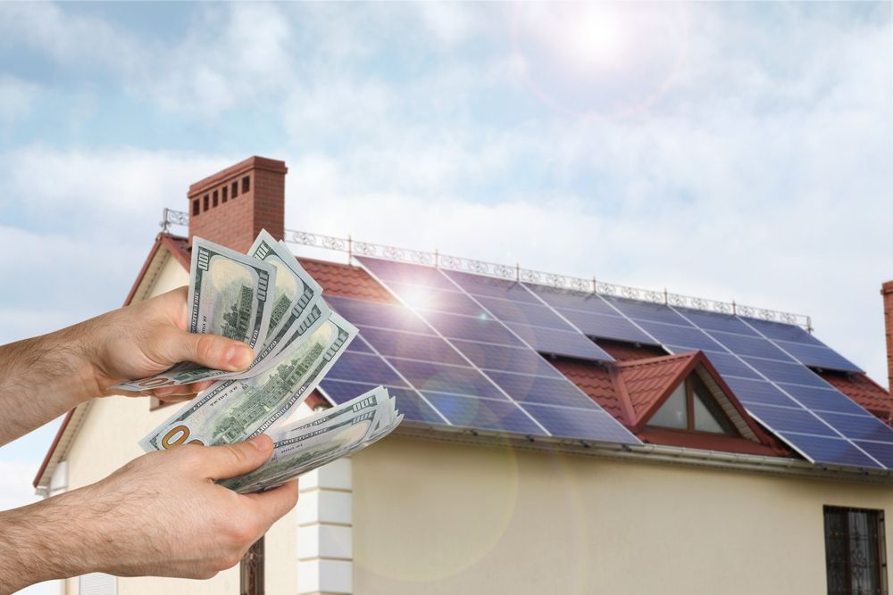 A person is holding money in front of a house with solar panels on the roof.