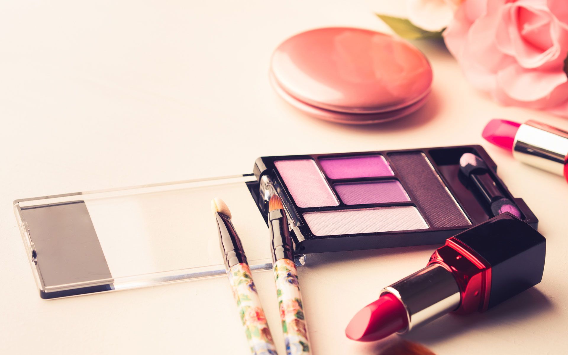 A lipstick , eyeshadow , brushes and a mirror are on a table.