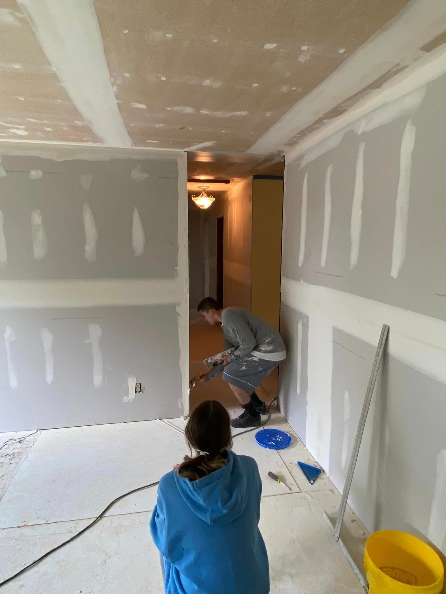 A woman in a blue hoodie is kneeling down in a room while a man works on the ceiling.