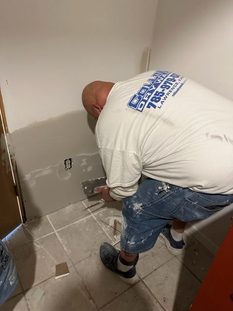 A man wearing a white shirt with a blue logo on the back is plastering a wall.