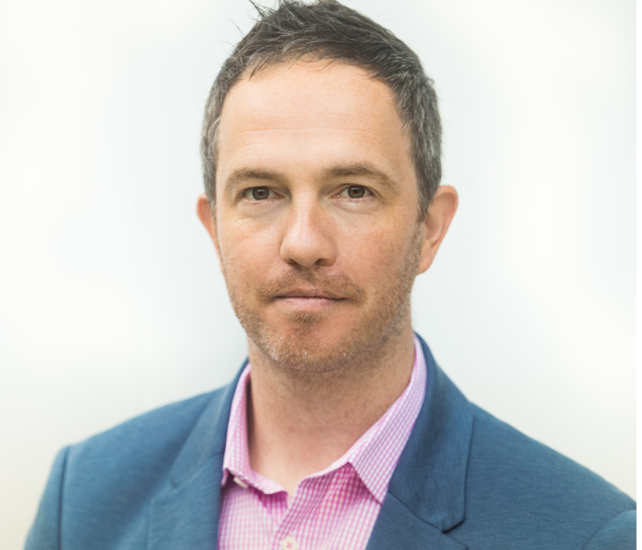 Man in blue blazer and pink shirt looking at camera, neutral expression.